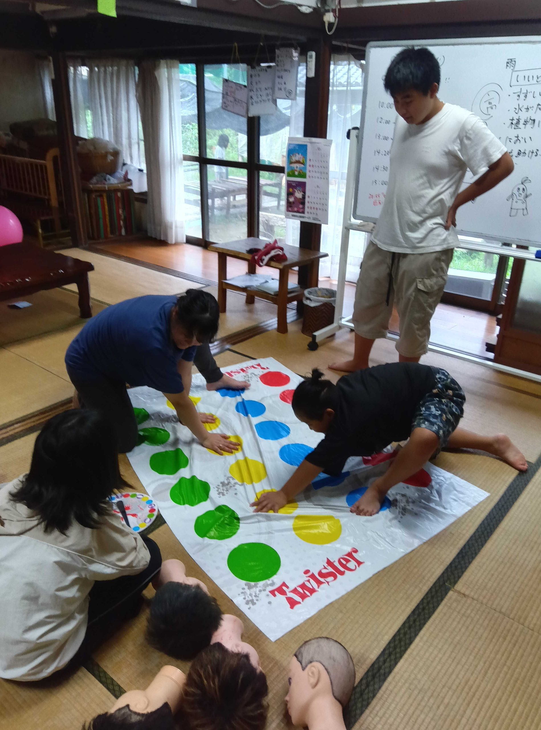ツイスターで盛り上がる子どもたちとスタッフ、奥には日課が書かれたホワイトボード