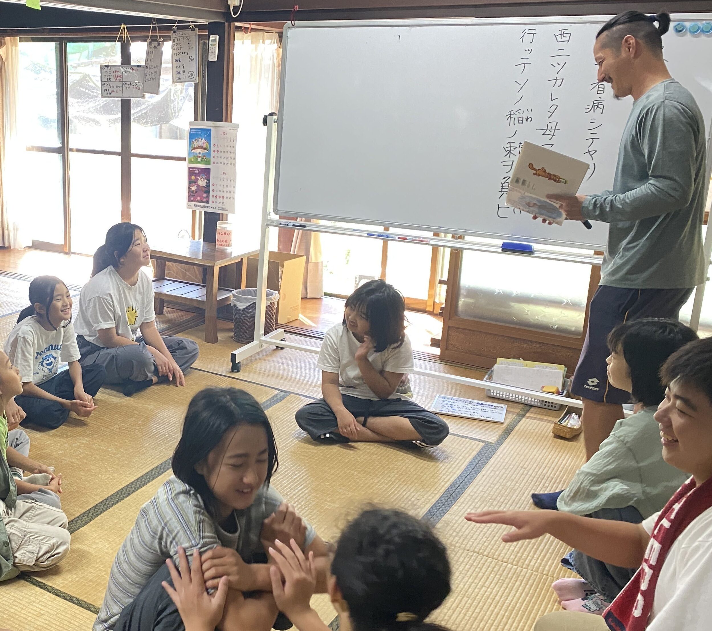 ホワイトボードに書いた「雨ニモマケズ」を読むスタッフと、輪になって座る子どもたち
