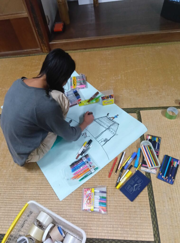 大きな紙に色ペンで家の絵を描く子