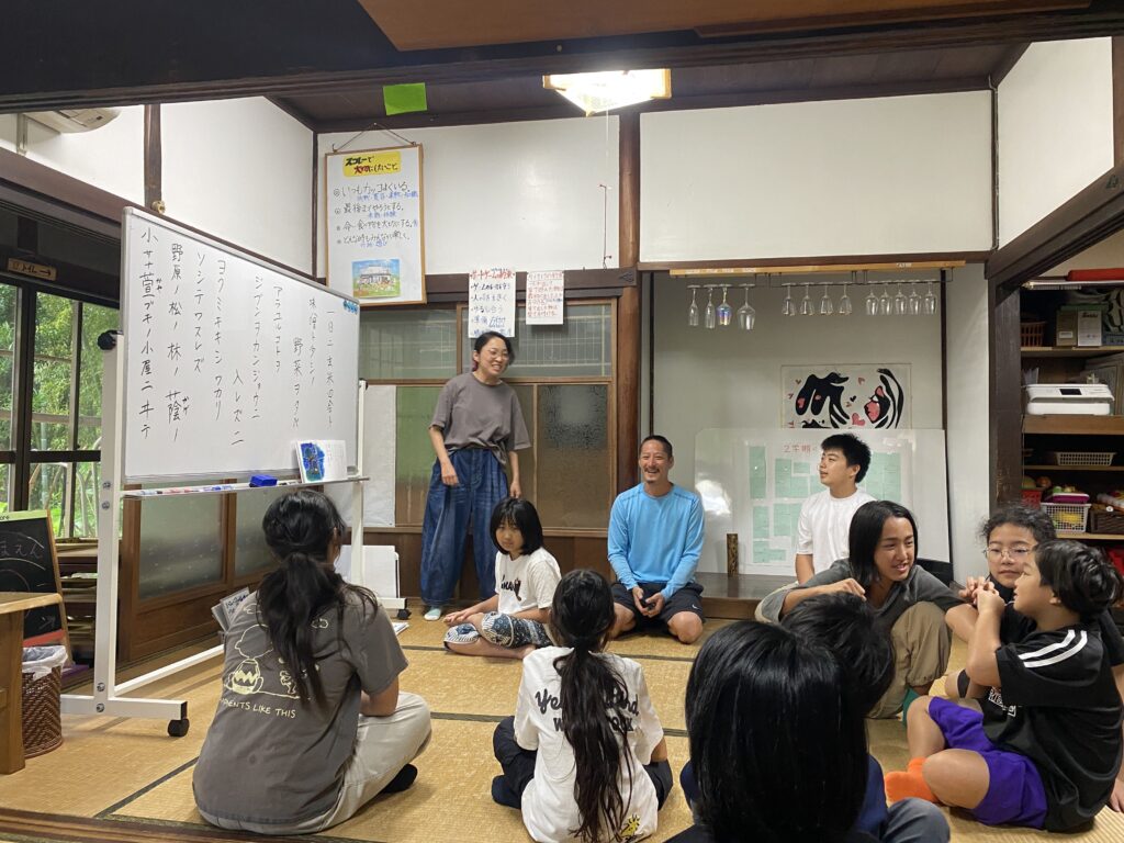 ホワイトボードに書かれた「雨ニモマケズ」を前に、輪になって座る子どもたちとスタッフ