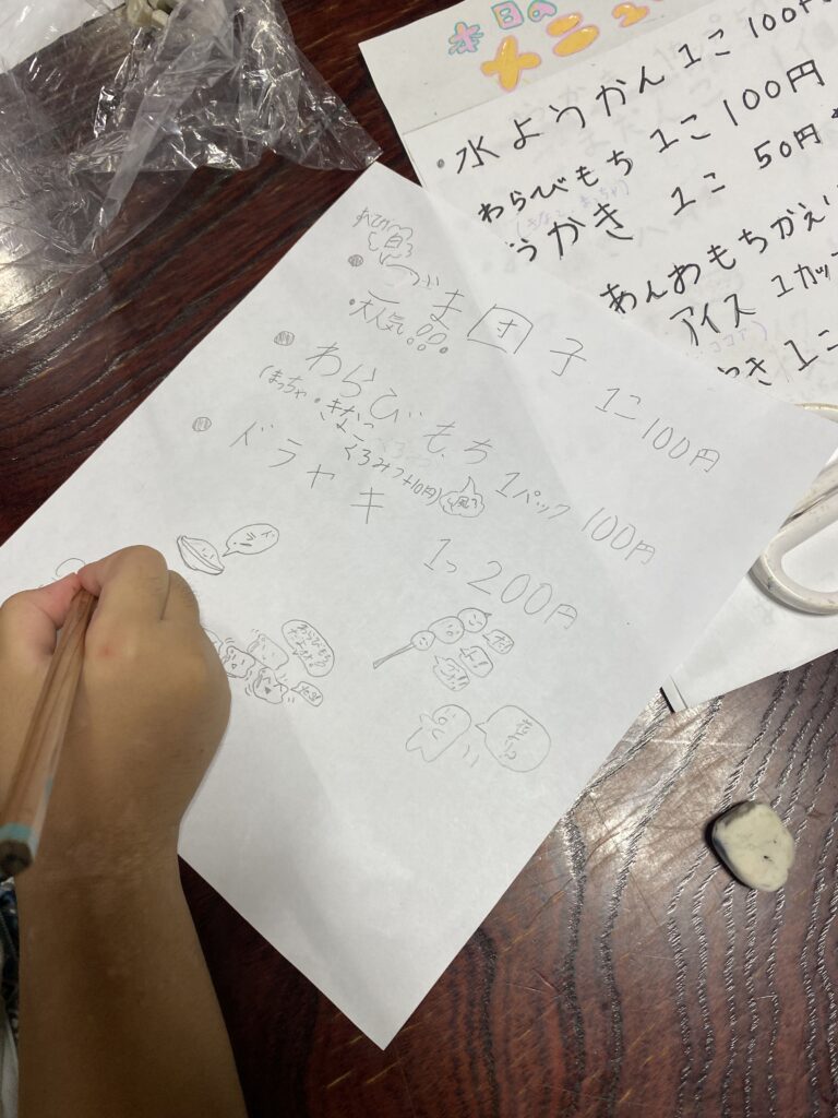 お菓子のメニューと値段を手書きで書き出す子どもの手元