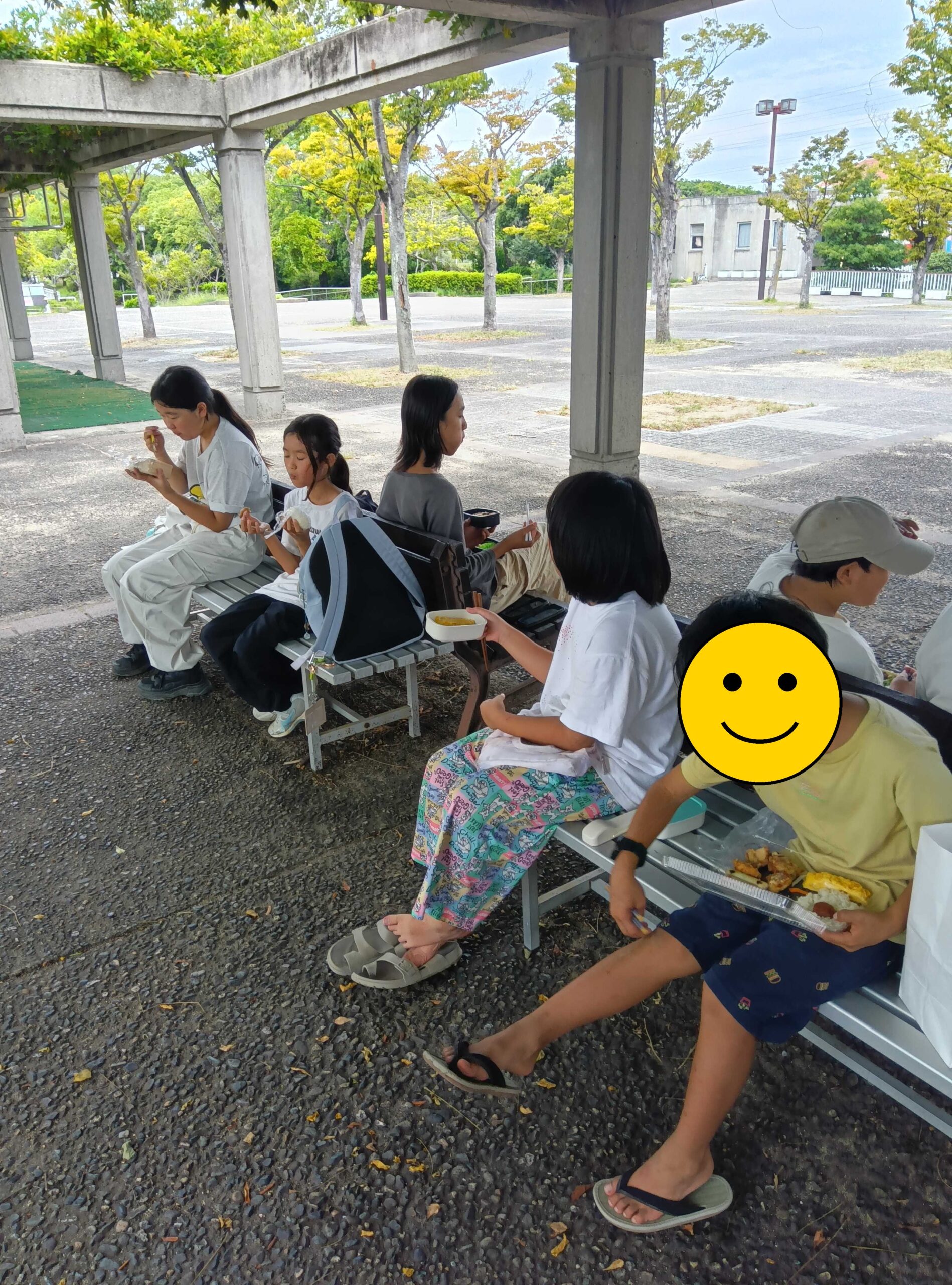 屋根付きのベンチでお弁当を食べる子どもたち(1人、顔にスタンプ加工)