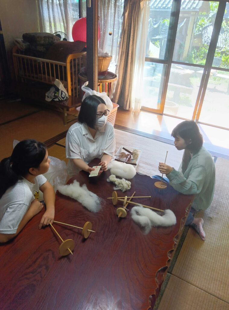 羊毛とスピンドルを広げた机を囲む子どもたちと大人