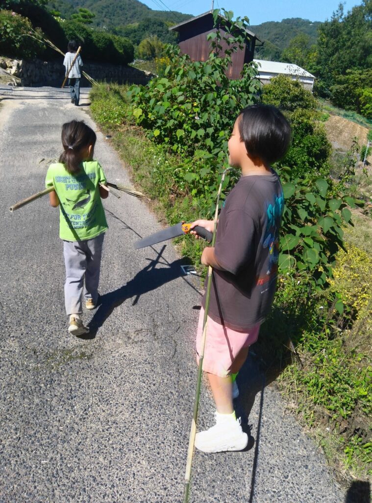竹の棒とのこぎりを持って道を歩く子どもたち