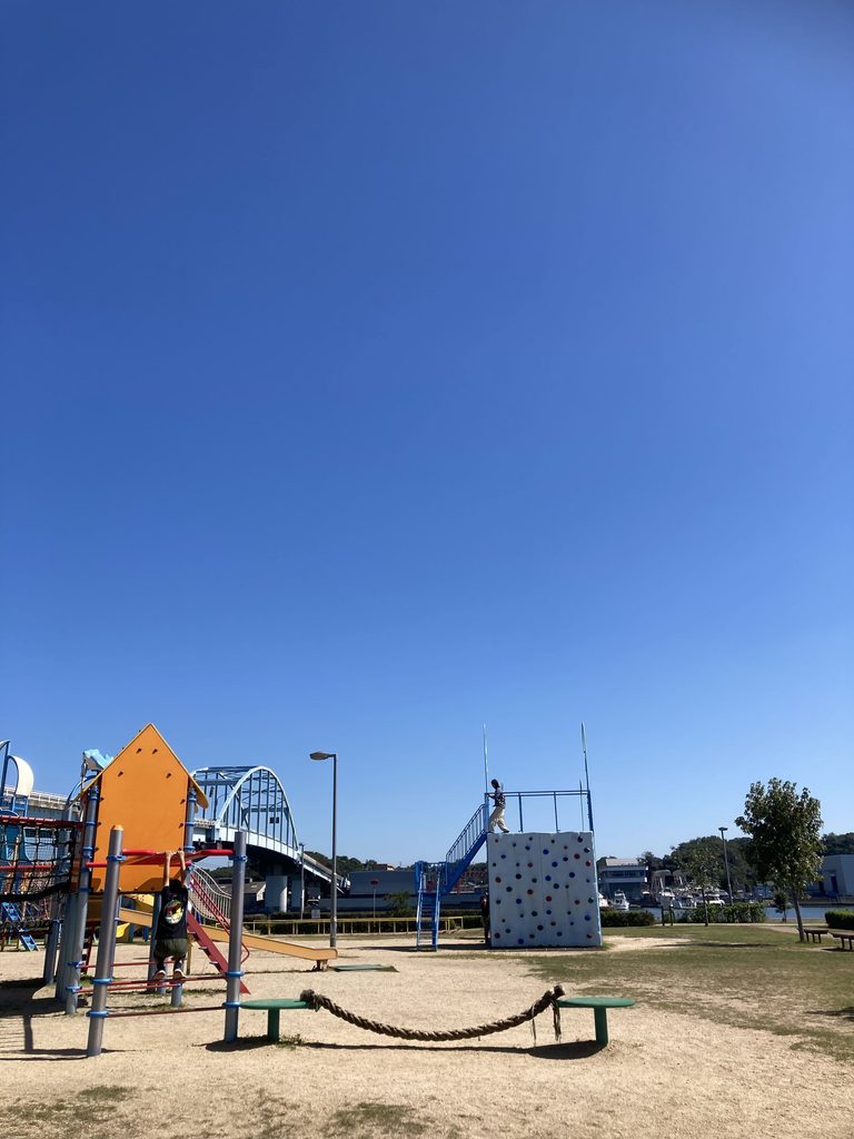 青空の下、橋の見える公園の遊具と、クライミングウォールに登る子ども