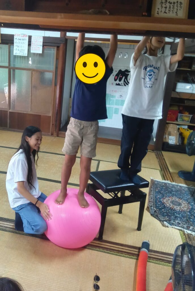 バランスボールの上に立つ子（顔にスタンプ加工）と、ボールを押さえて支える子
