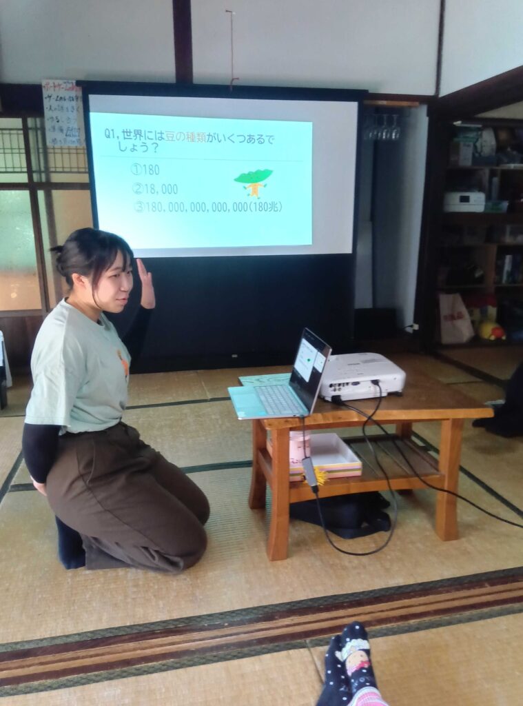 「世界には豆の種類がいくつあるでしょう？」というスライドを映して説明する大学生ボランティア