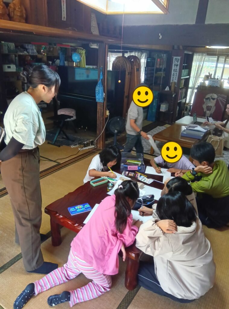 ちゃぶ台を囲んで絵を描く子どもたちと、それを見守る大学生ボランティア（顔にスタンプ加工）