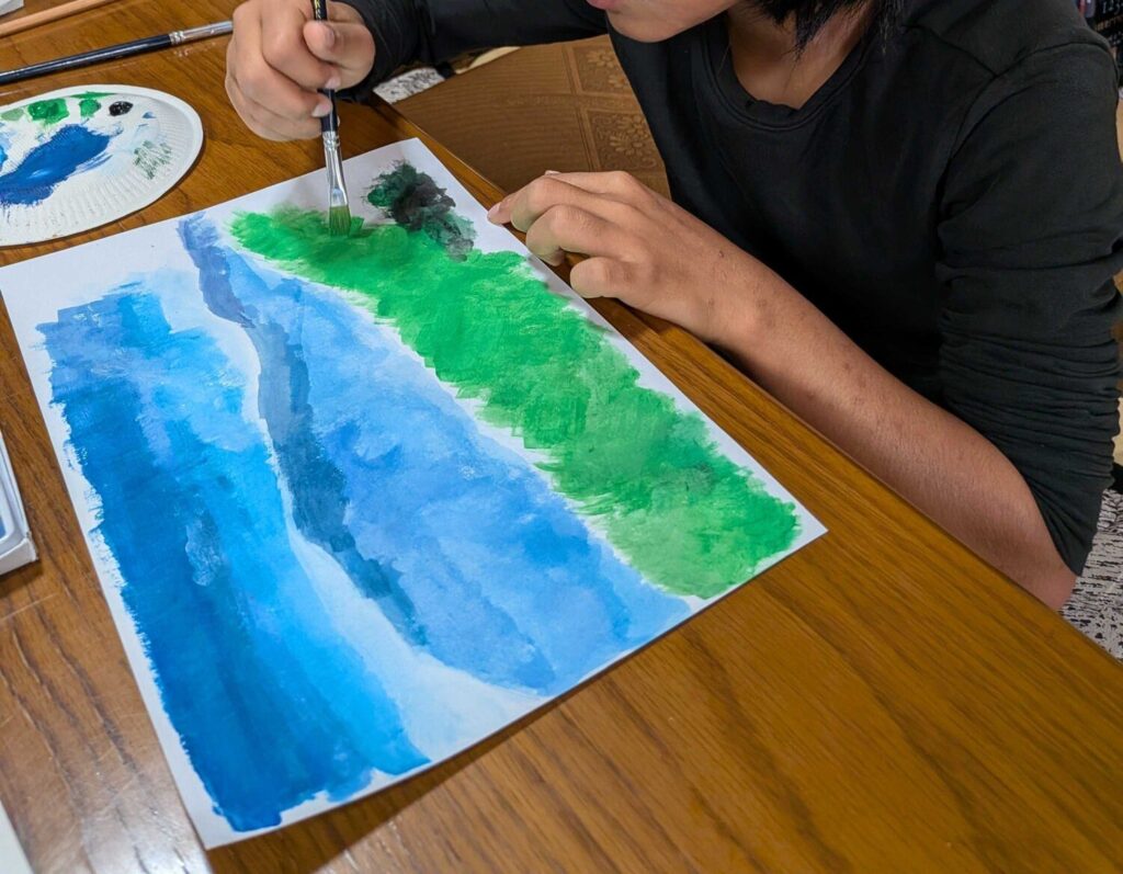 青と緑の絵の具で、画用紙に絵を描く子ども