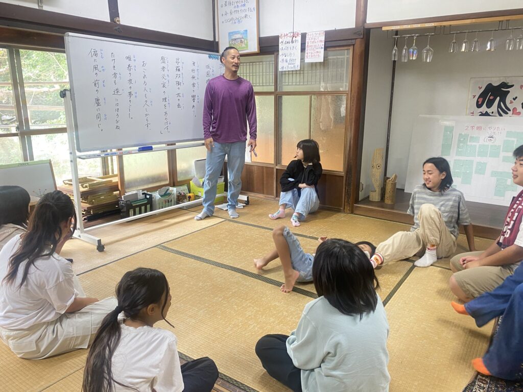 平家物語の冒頭が書かれたホワイトボードの前で、輪になって座る子どもたちとスタッフ