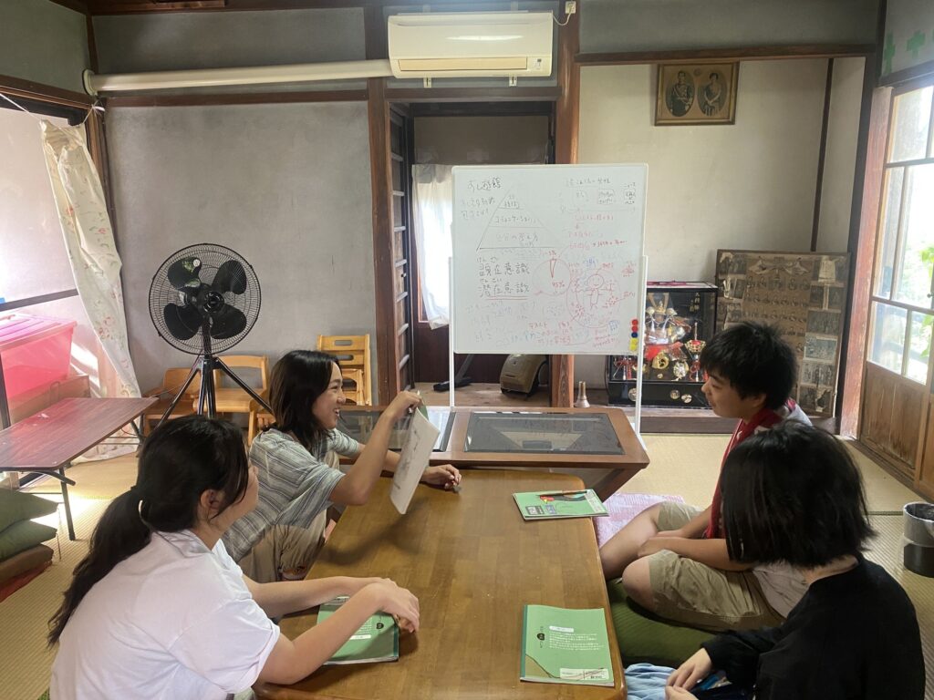 ホワイトボードのある部屋で、机を囲んで話を聞く中学部の子どもたち
