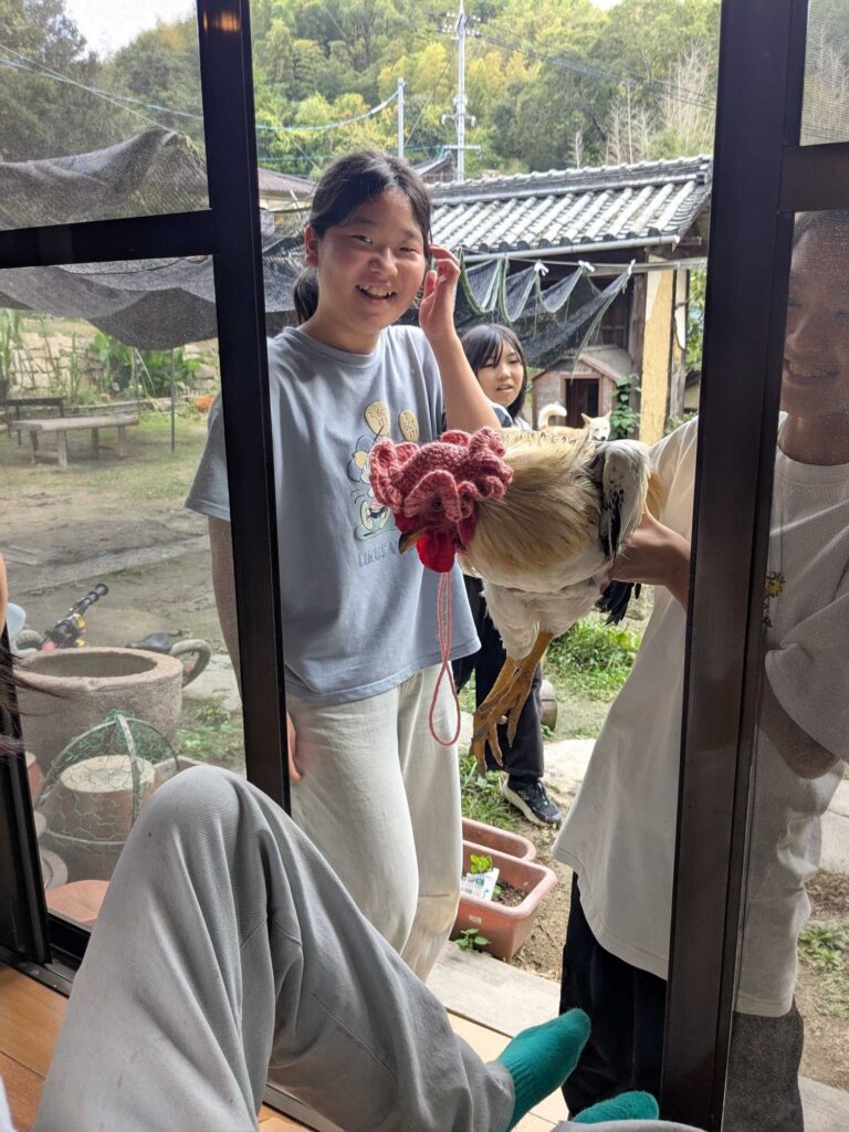 毛糸の帽子をかぶったニワトリを抱えて、窓の外に集まる子どもたち