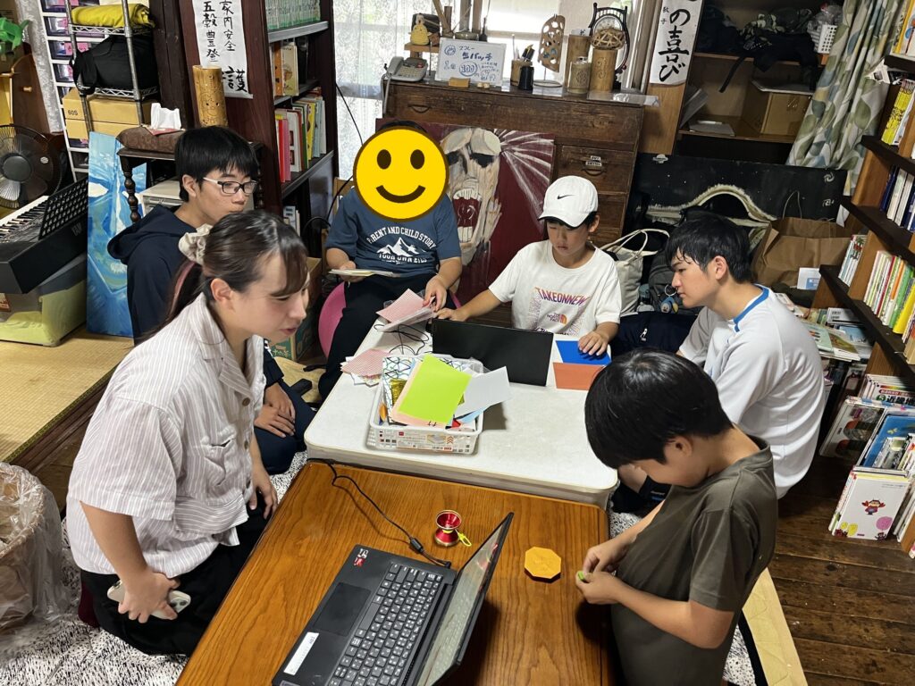 高校生も一緒に、折り紙とパソコンの机を囲む様子
