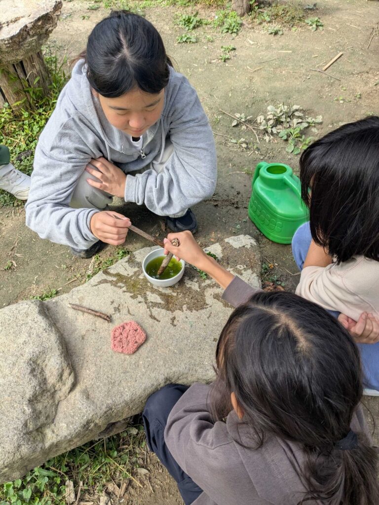 器に入った緑色の水を、枝でかき混ぜる子どもたち